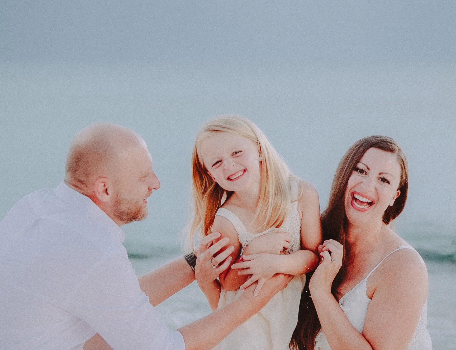 Parents laughing with their daughter on the beach under soft grey clouds in Grand Cayman