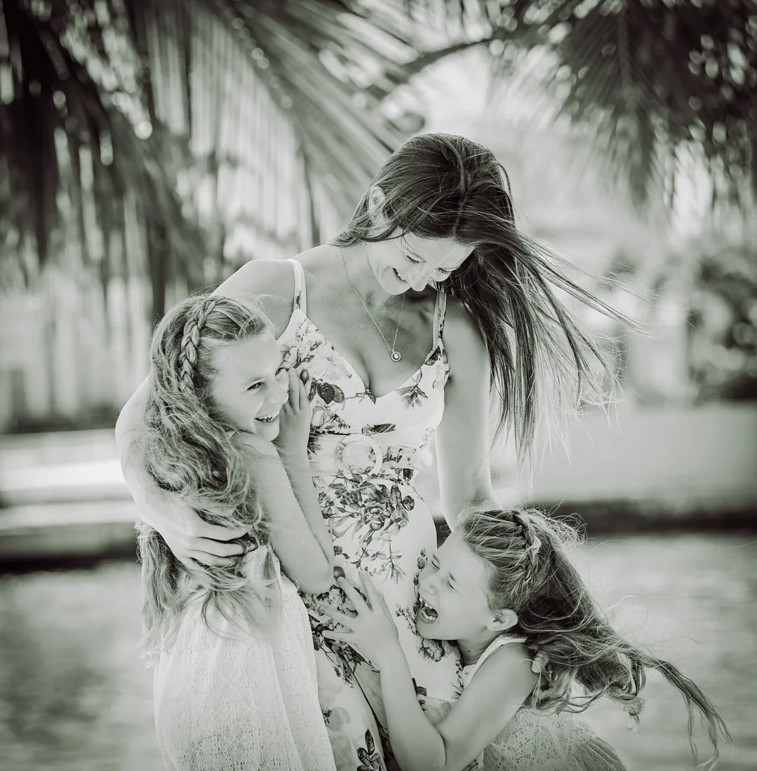 Mother in a floral dress laughing as her two daughters hug her beneath palm fronds in Grand Cayman
