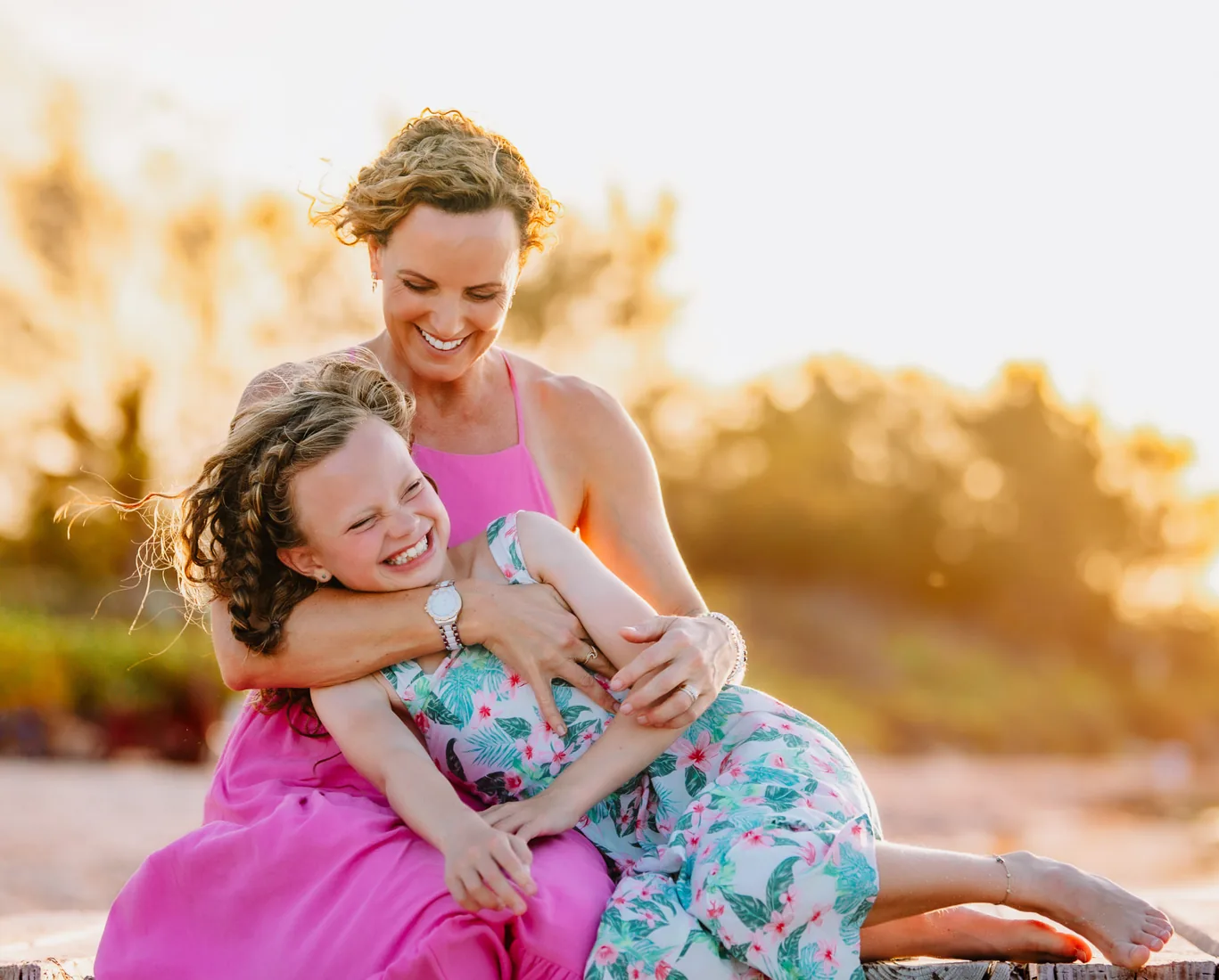 Mother cuddling her laughing daughter as they sit together on the beach at sunset