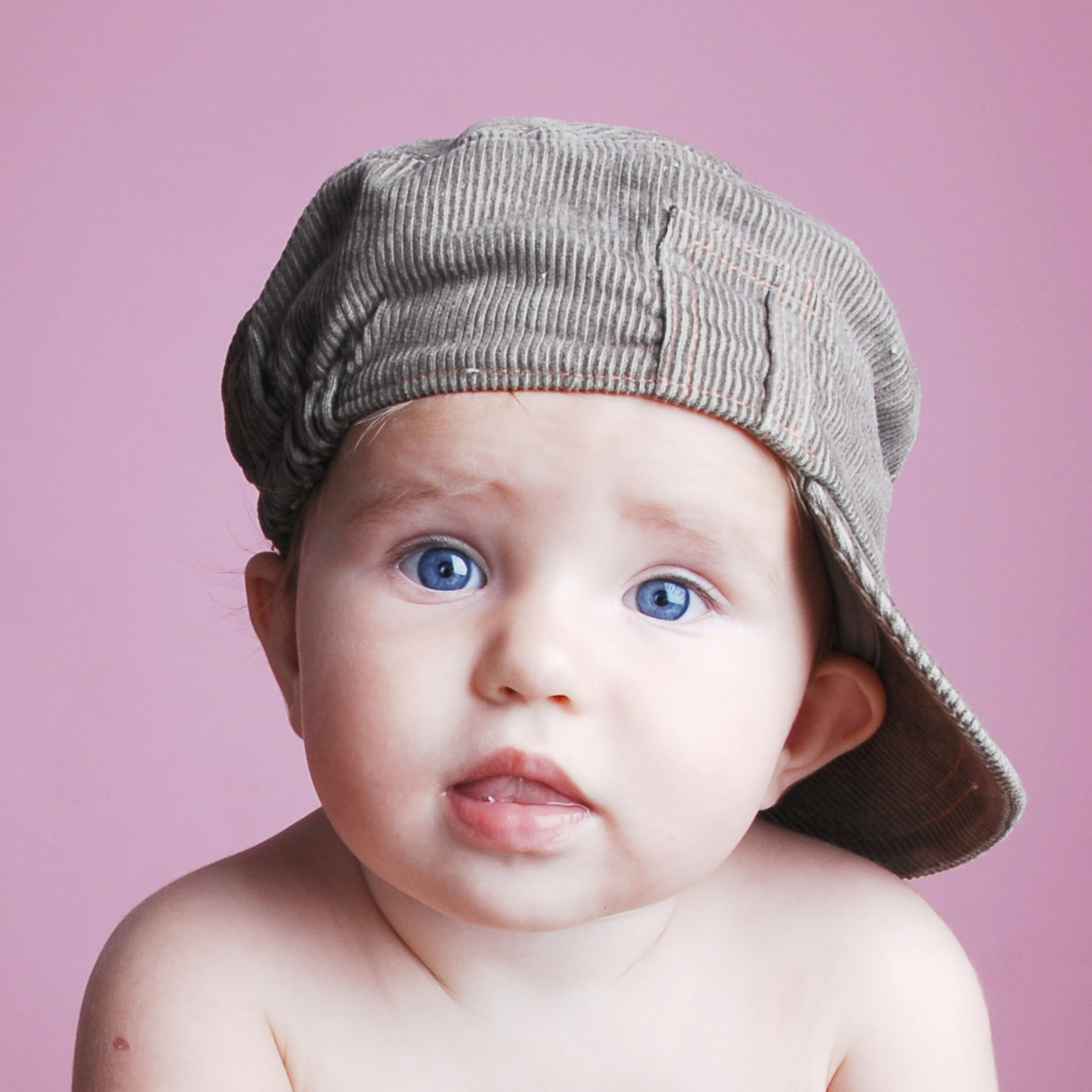 Baby with bright blue eyes wearing a soft cap against a pink backdrop