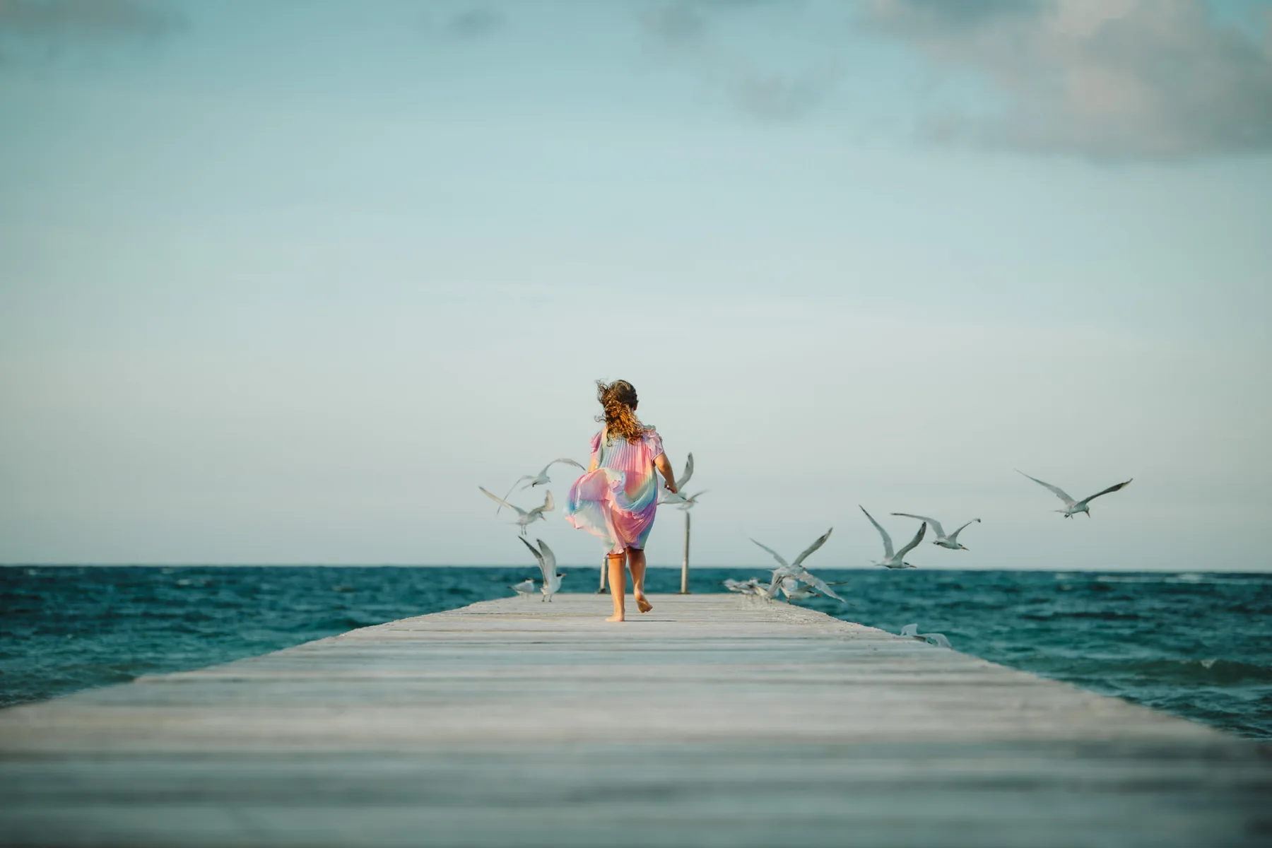 Girl running along a wooden dock as seabirds take flight over the ocean in Grand Cayman