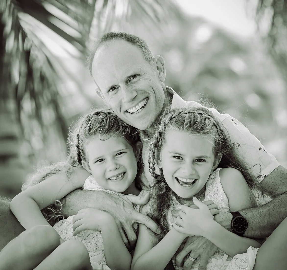 Father embracing his two laughing daughters outdoors in soft island light in Grand Cayman
