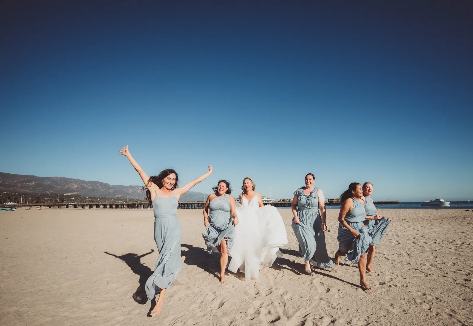 Bride and five bridesmaids running barefoot across the sand with the ocean and a pier behind them