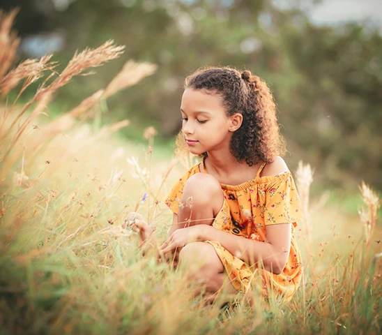 Girl in a mustard sundress crouching in a golden meadow holding a grass stem in Grand Cayman