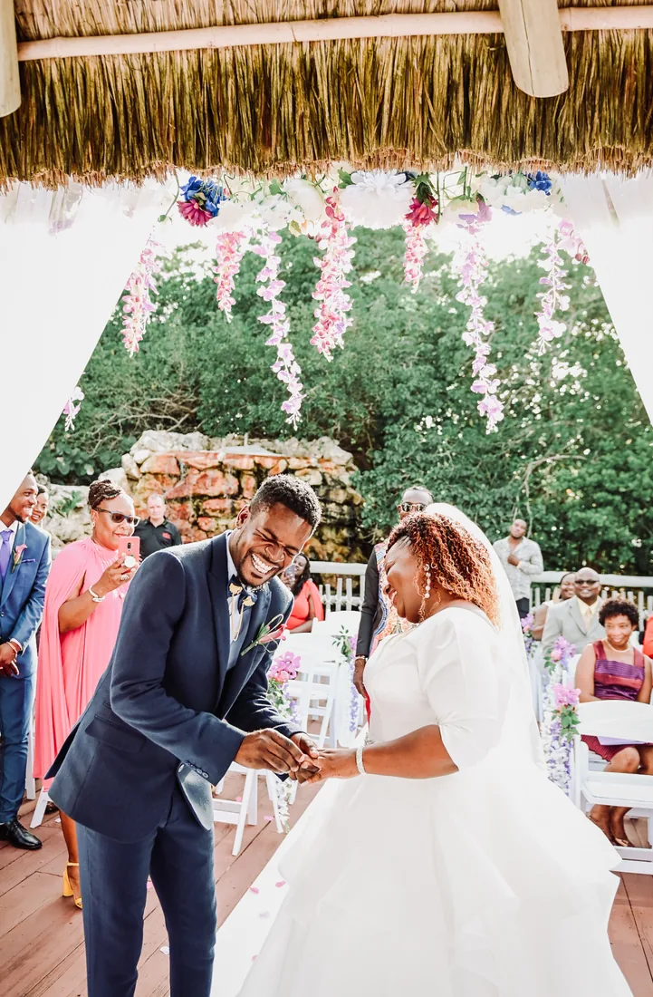 Groom slipping the ring onto the bride's hand beneath a flower-draped canopy as guests look on in Grand Cayman