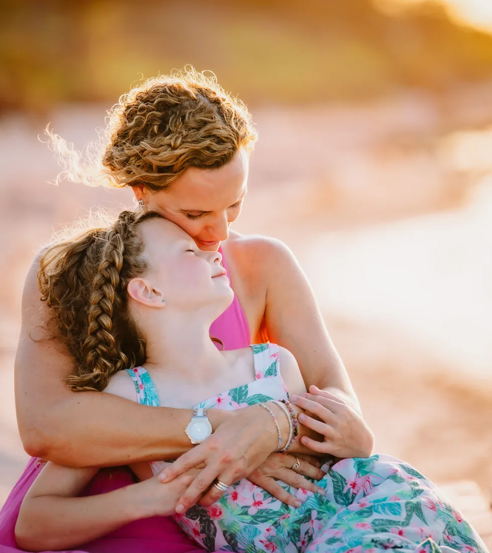 Mother embracing her daughter who rests in her lap on the sandy beach at sunset