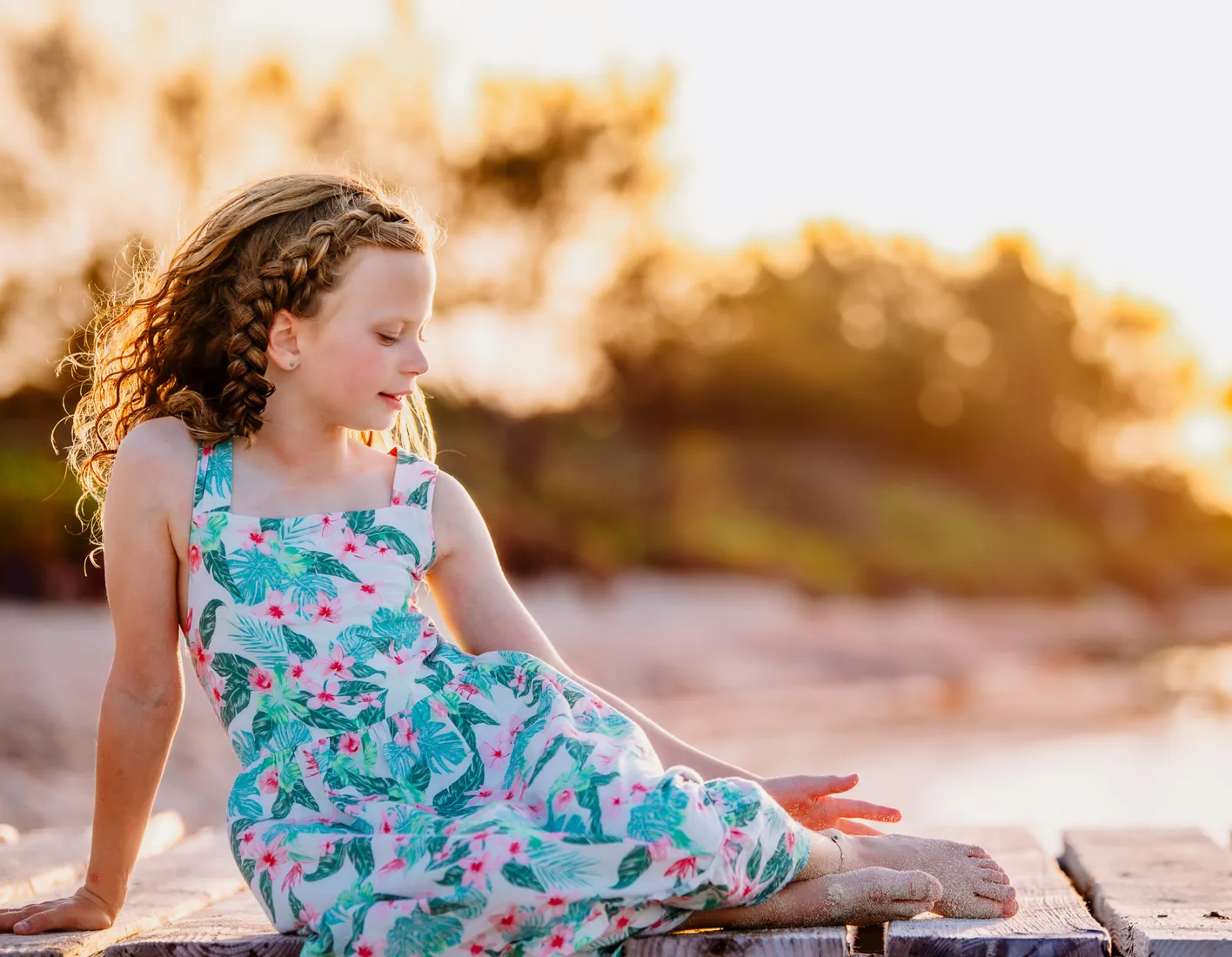 Girl in a palm-leaf and flower sundress sitting on a weathered wooden dock at golden hour in Grand Cayman